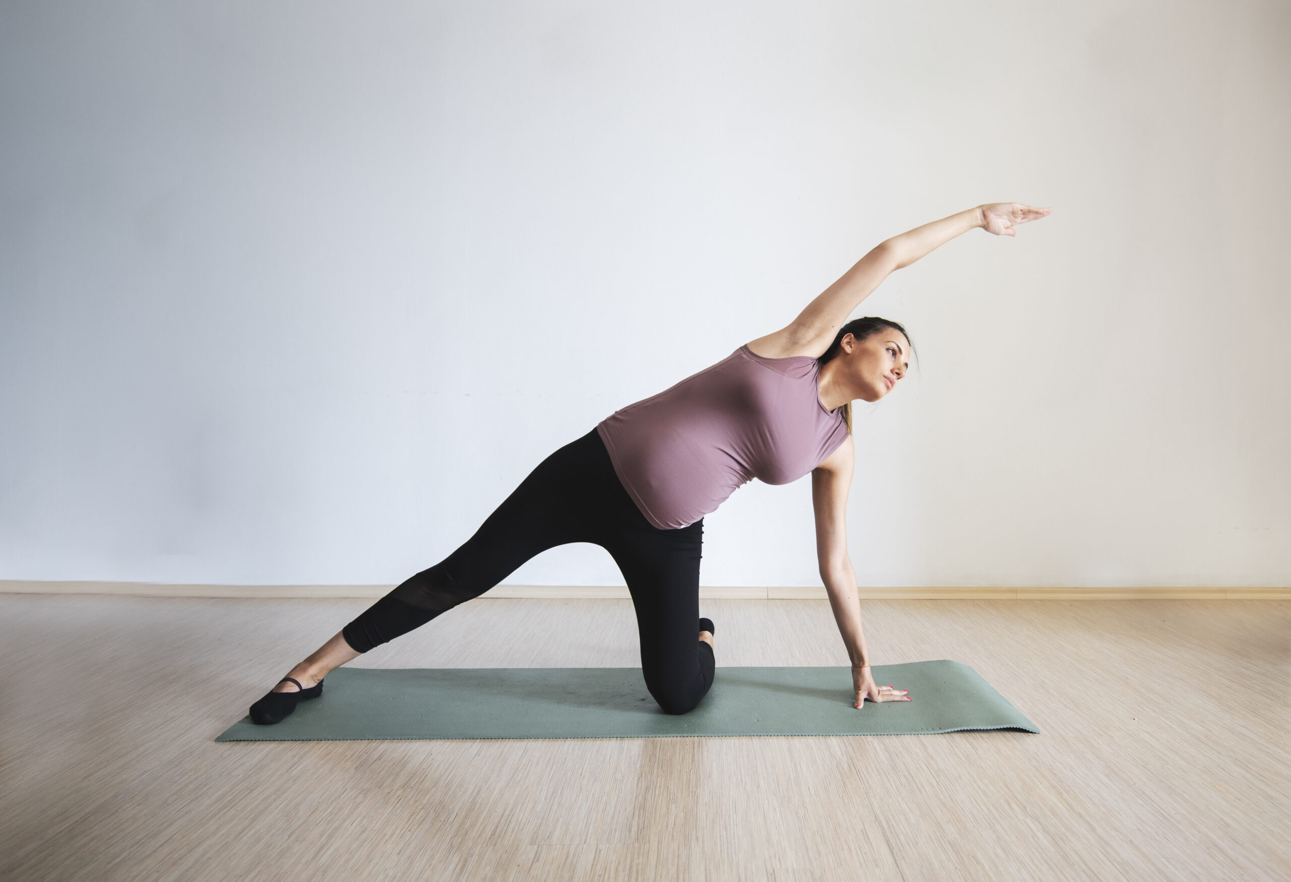 Schwangere Frau in Sportsachen, die auf einer Yogamatte kniet und den Arm lang über den Kopf streckt