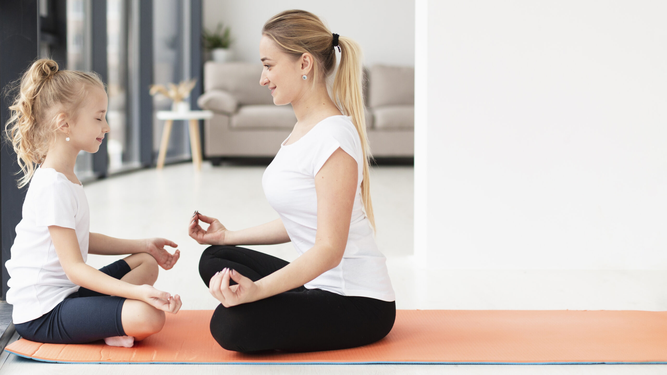 Mutter und Tochter sitzen gemeinsam auf einer Yogamatte sich gegenüber im Schneidersitz und lächeln